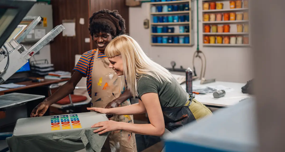 Two print-shop workers lifting a freshly pressed full-color print on a T-shirt at a heat press